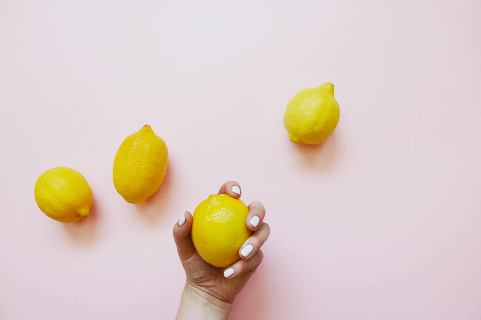 A hand holding a lemon on a soft pink background surrounded by additional lemons, symbolizing consistent yet varied pleasure.
