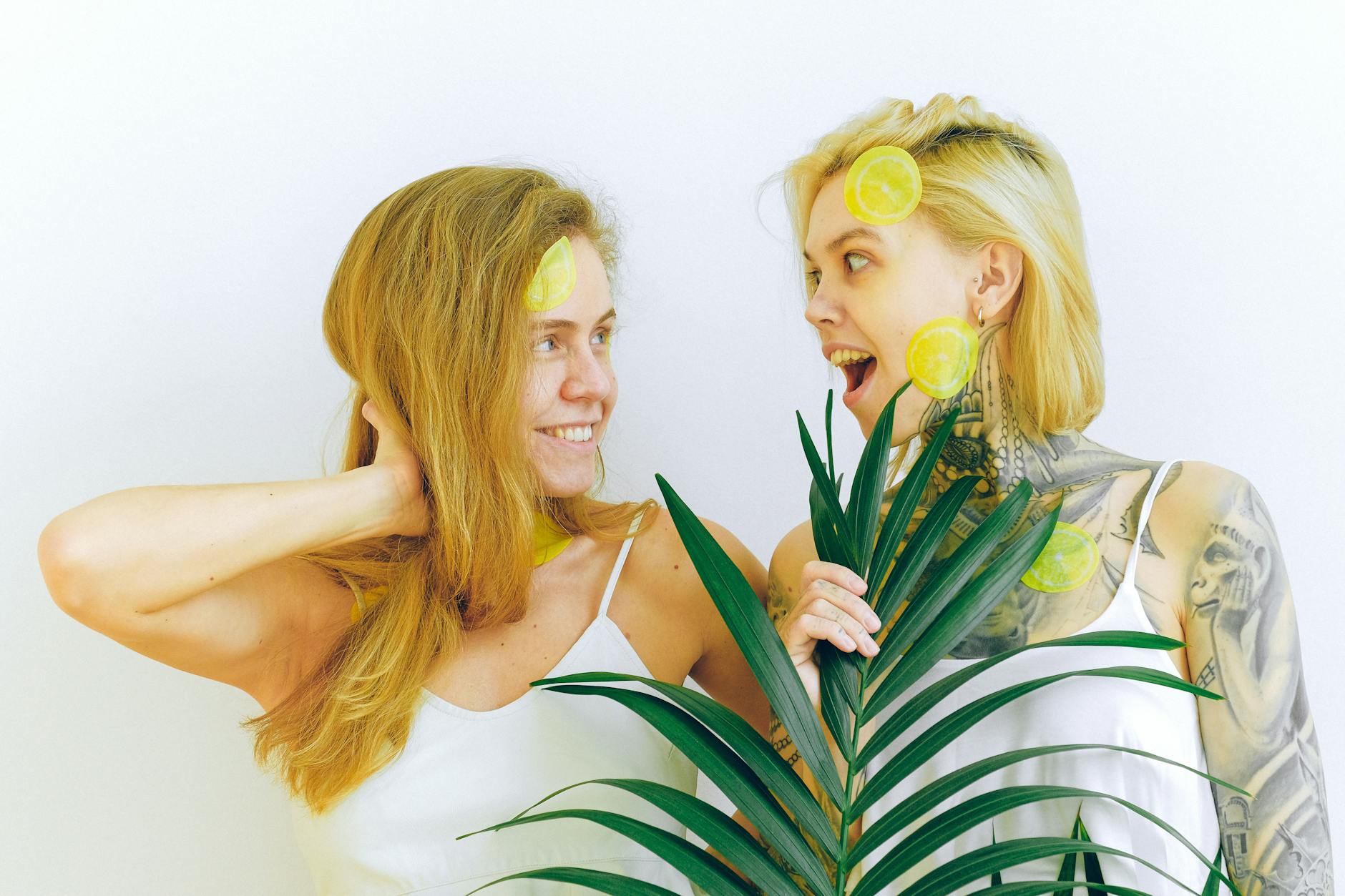 Two women smiling and laughing together, holding lemon slices in front of a vibrant green plant, conveying joy and ease.